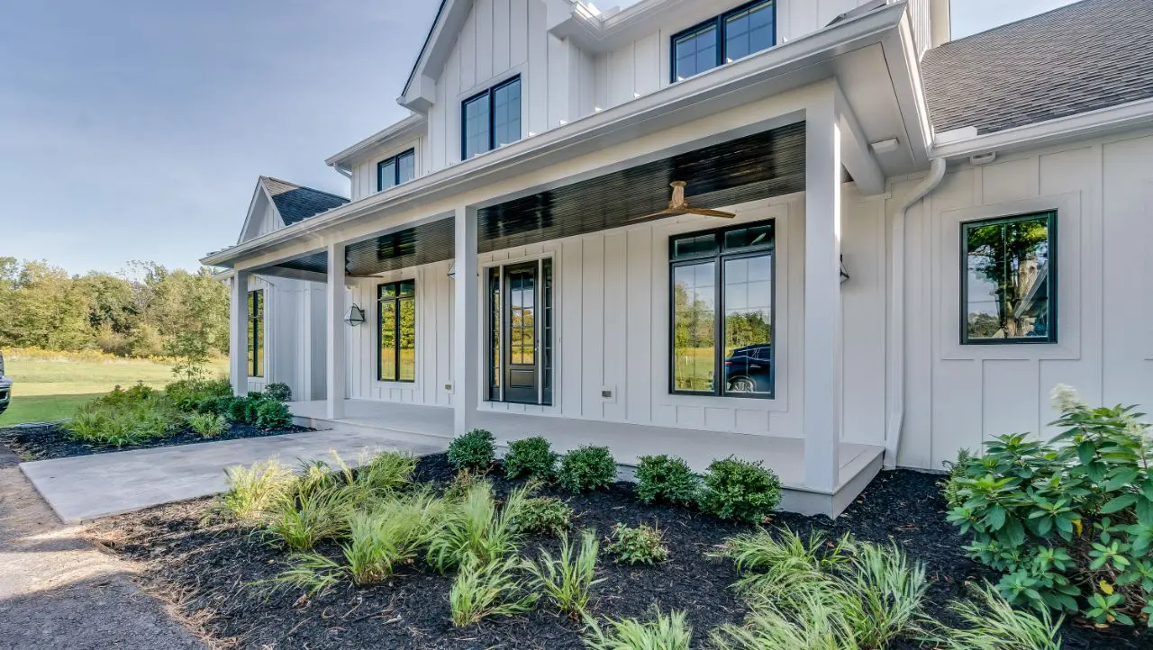 This custom-built, white modern farmhouse features a sleek black roof and expansive windows, nestled in a beautifully landscaped front yard adorned with lush shrubs and plants.