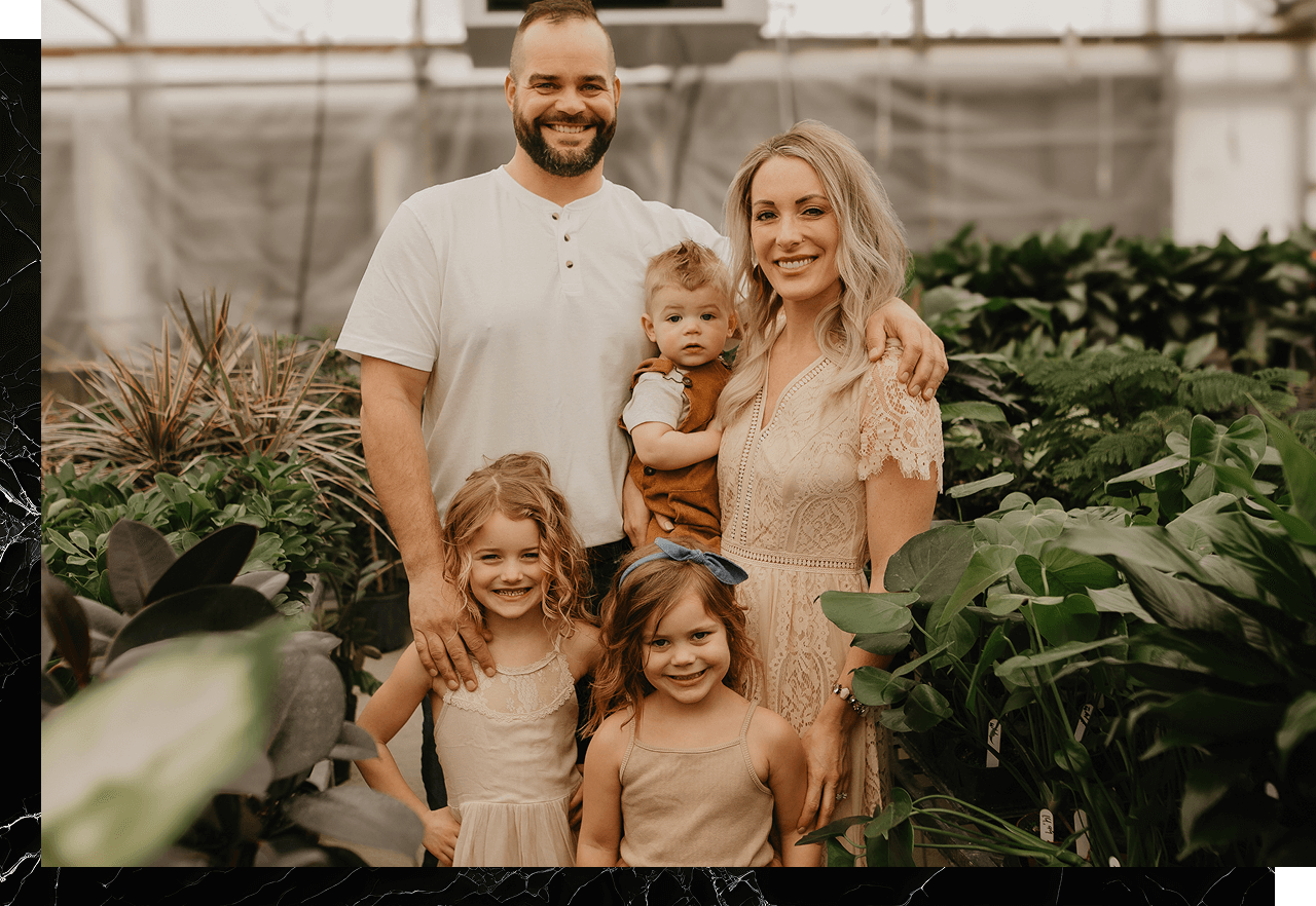 Amid the verdant wonders of the greenhouse, a family of five stands together, posing among leafy plants. Behind their two children, the parents proudly hold a baby. Just as nurturing plants takes time, they wonder about the future and how much it might cost to build a house for their growing family.